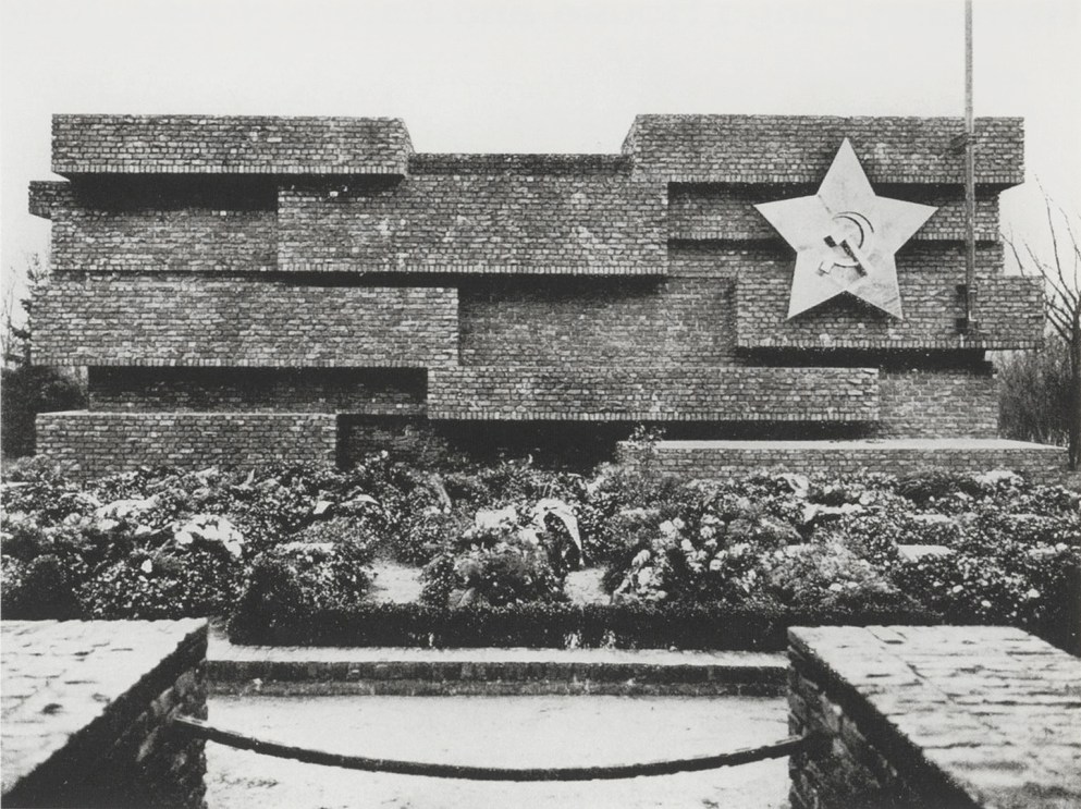 memorial to the november revolution was a horizontally oriented solid jenga structure of unaligned and asymmetrical blocks, constructed of brick salvaged from buildings damaged in the revolution, or the walls against which anti-fascists were shot. a large five pointed star with a hammer and sickle on the upper right section of the memorial had a flagpole attached next to it. a low border of flowers in the berlin cemetery where the memorial was built sits behind a gravel sidewalk, and a chained, low gateway. designed by mies van der rohe and destroyed by nazis