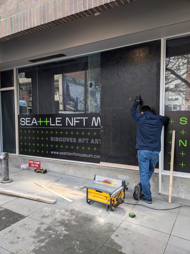 a photo of an art handler installing a sheet of black-painted plywood across the smashed window of the seattle nft museum, with a small portable table saw on the sidewalk, and sawdust and tools and some dimensional lumber and a green-striped tape measure, for atmosphere, as tweeted jan 14, 2022 by socialist suits