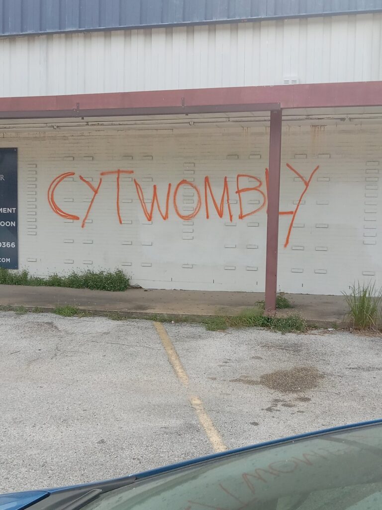 a photo across the windshield of a car of a cy twombly tag in orange spray paint on the dingy painted brick facade of the long-ago closed sand dollar thrift shop in houston, as tweeted by @buffalosean