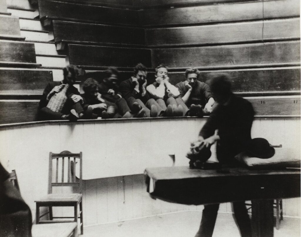 photo of the operating theater at jefferson medical college in philadelphia, where six young goofballs wearing old-timey fits have their feet up on the railing, their elbows on their knees, and their hands under their chins as they watch a blurry (thus, moving, because it's a 19th century photo) male figure pretend to examine the crap out of a skull on the operating table. the viewer on the far left is holding a wicker-encased jug of something, and the guy next to him has a cup, so it's a party. there's a chair sitting empty in the lower left corner, and the upper half of the photo is empty bleacher seats.