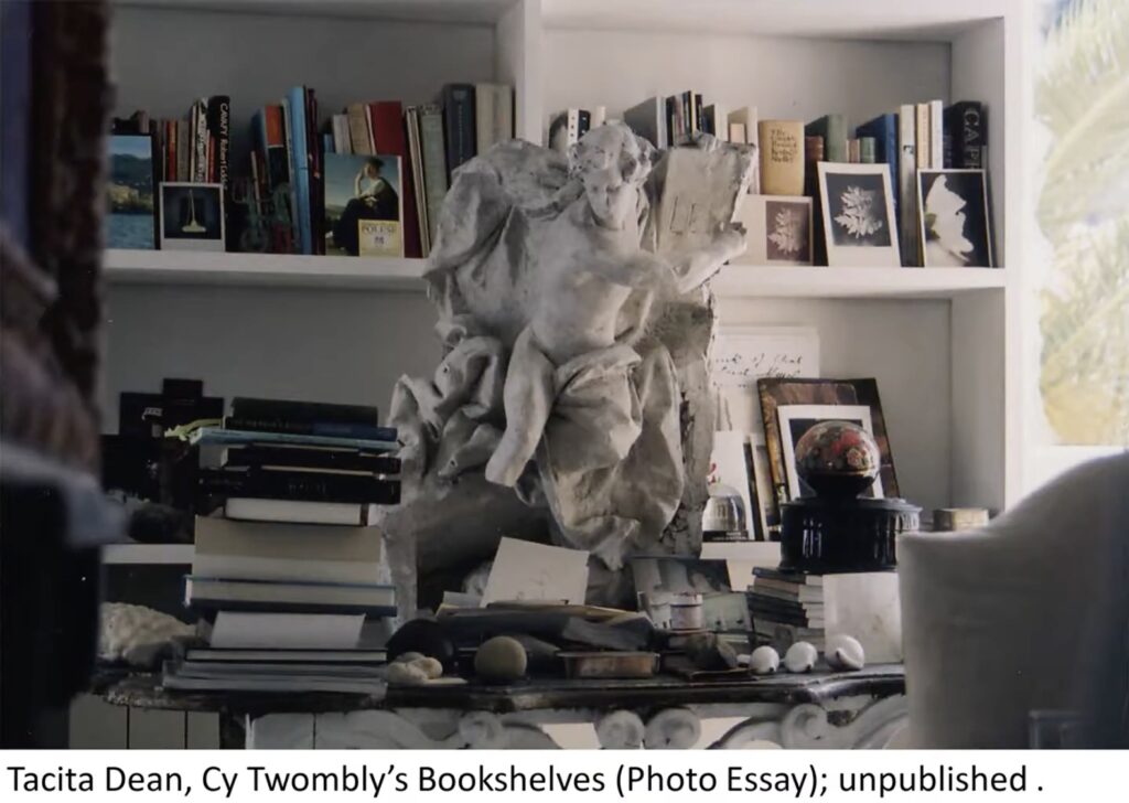 a plaster or white painted terra cotta statue of a male nude figure twisting to hold a tablet while billows of drapery surround him sits on a table in front of a wall of books in cy twombly's living room slash bedroom in gaeta. tacita dean made this photo in 2008 but did not publish it, though poet mary jacobus used it in a presentation, from whence this screengrab was made in 2023, in order to identify the statue.