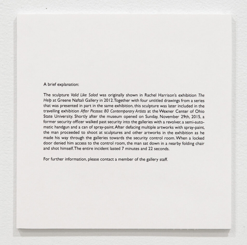sign on a gallery wall with a paragraph of text explaining why this sculpture has a bullet hole in it now: tl;dr it was shot while on display at the wexner center in columbus ohio.