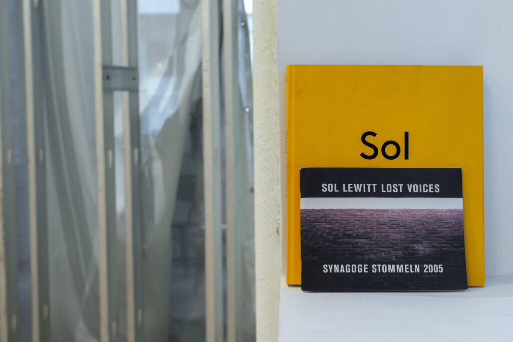 A color photograph of a windowsill with a larger yellow book and a small black book. The yellow book reads "Sol" in black text. The front book reads "Sol Lewitt Lost Voices Synagogue Stommeln 2005" in white text. To the left of the windowsill is a piece of semitransparent fabric hung from grey metal scaffolding.