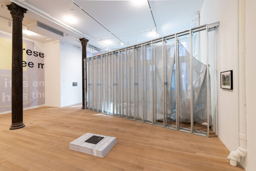 A color photograph of a gallery space. A large piece of grey and yellow vinyl hangs from the wall on the left, perpendicular to diagonal temporary wall of aluminum studs with semi-transparent fabric on one side. A stack of grey and white posters are placed on the ground directly in front of the unfinished wall.