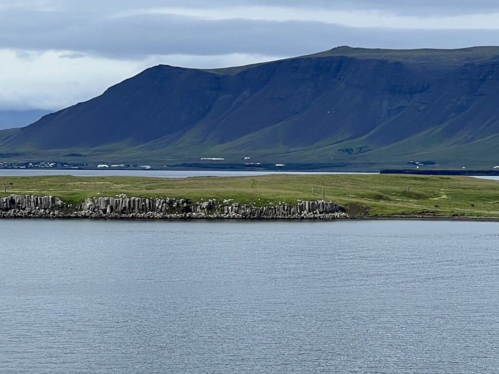 videy island in the middle ground, water in the lower foreground, and a dark grey and green mountain under cloudy sky, as photographed from the deck of a ship