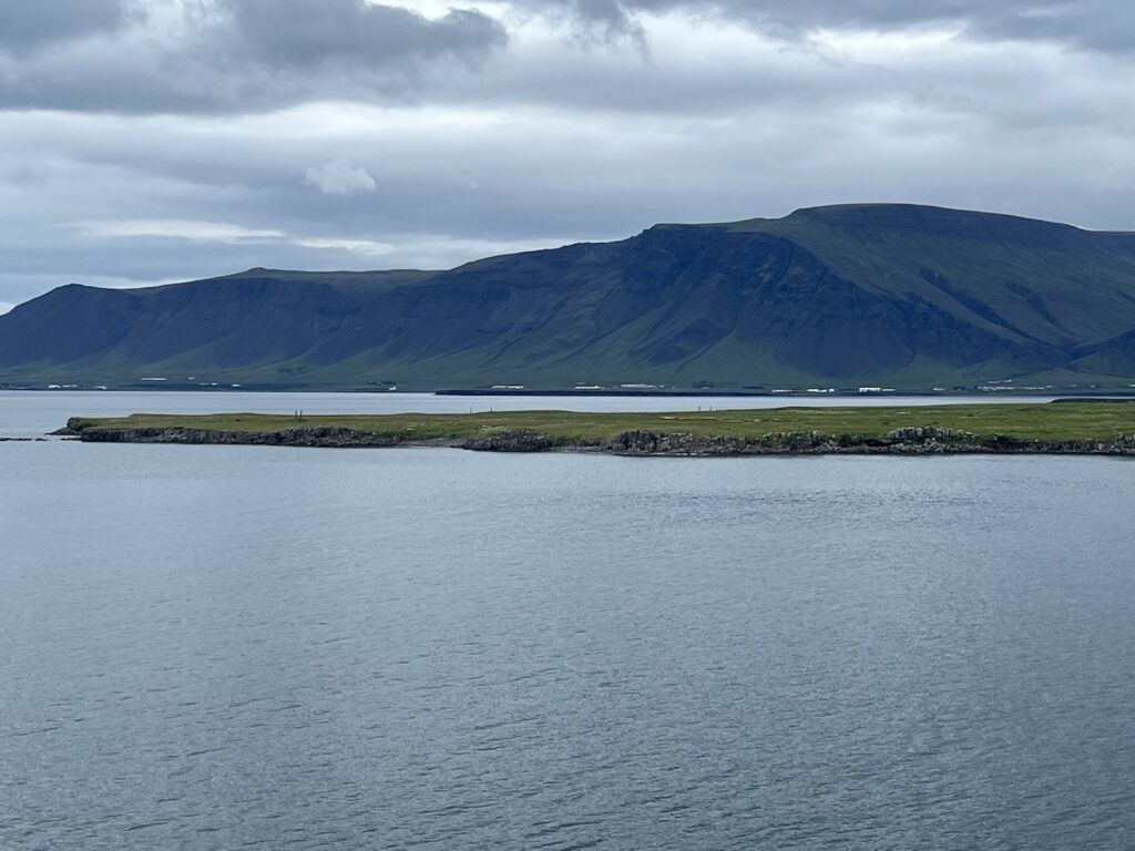 videy island very flat in the center, with richard serra's tall stone monoliths barely peeking above the landscape. the sea in front and a mountain in back, taken from a ship.