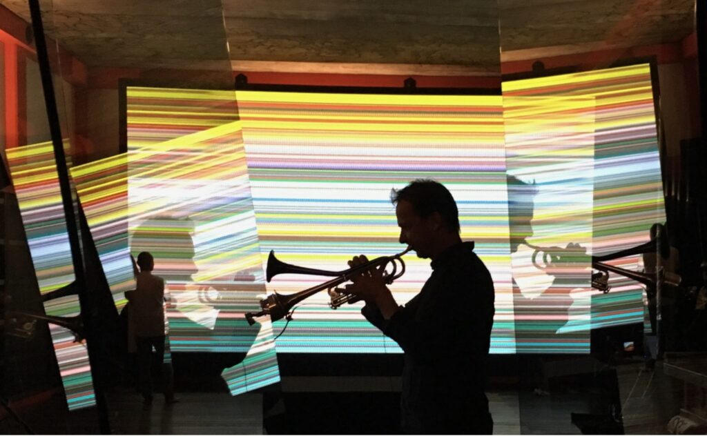 trumpeter marco blaauw silhouetted against gerhard richter and corinna belz' digital animated film, moving picture (946-3) kyoto version, which at this moment is a multicolored stack of thin horizontal lines, is reflected in angled panes of glass on the right and left, because the photographer took the shot through richter's sculpture, 8 panes, which was also exhibited in kyoto at the same gig in 2019.