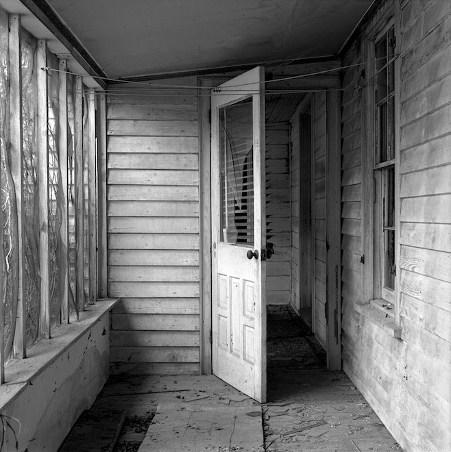 david simonton's black and white photo of a white painted wood door with an intact glass window left ajar on what seems to be the screened in rear or side porch of a worn, possibly abandoned clapboard house. the screen netting sags between slats on the left, the worn off paint on the right, some lumber and dirt and light debris on the porch floor, and a couple of laundry lines suspended across the space up top. taken, simonton says, between 1993 and 2011, in nahunta, nc