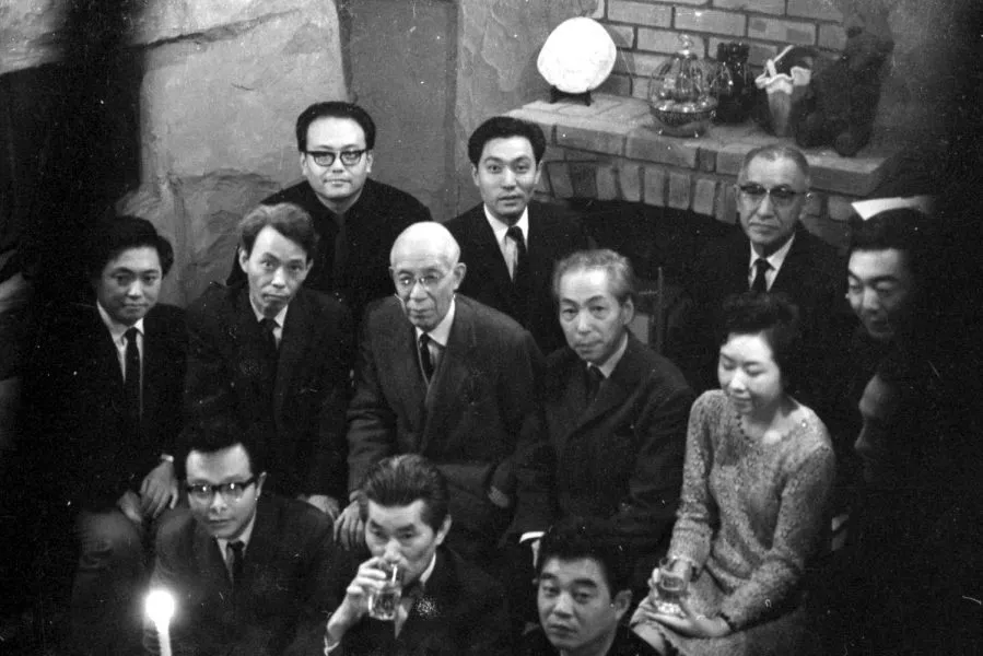 a group of japanese men in suits and one woman seated in a rock walled bar in harajuku in 1967, celebrating the completion of takiguchi shuzo's proof of poetic experiments: 1927-1937. looks like quite a party