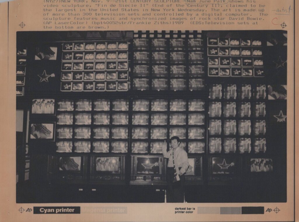 1989 ap photo of artist nam june paik standing in front of his sculpture fin de siecle ii at the whitney museum. it is a wall of over 300 television sets in various sizes and grid configurations showing seven different channels of programming, but the caption only says its david bowie. popped up on ebay, then disappeared oh well