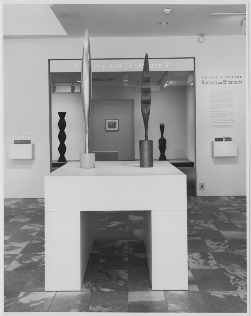 a black and white installation photo of scott burton's exhibition of brancusi sculptures at the museum of modern art in 1989 depicts the entrance of the exhibit, where a thick pedestal sculpture by burton in the shape of a squared off, inverted u has two brancusi bird sculptures, removed from their original bases. beyond is a gallery with more brancusis; on the wall near the entrance are two small brochure holders, also created by burton. the distinctive thick grey and white grain of cesar pelli's moma circulation spaces is making me wish we could turn back the clock, but then I remember 1989, and maybe it's not a wistfulness for the past that makes me want to reverse time, but the dread of the immediate future. anyway, photo credit by mali olatunji