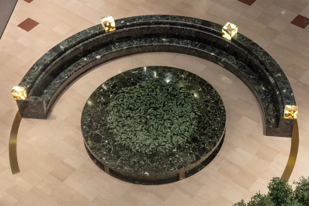 a semi circular green granite bench with a matching circular green granite planter in the center filled with leafy plants was originally installed in the atrium of the equitable building in manhattan, and is photographed from above. it is by scott burton and has been dismantled, as the ny times discussed at length in an october 2024 article from whence this photo was ganked.