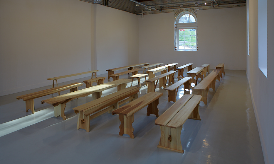 a white walled gallery with light grey floor is filled with light from three arched windows, which shines on six rows of benches, all made by francis cape from poplar, in designs carefully documented from various utopian communities in the usa. the exhibition at arcadia university took place in 2012.