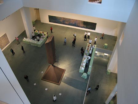 gothamist photo from a moma catwalk facing down onto the atrium, which is being set up for a party/preview, with drink stations and cater waiters milling about. the green grey slate floor dominates. barnett newman's bronze sculpture, broken obelisk, an obelisk perched upside down on the tip of a pyramid, is at the atrium center. on the back wall, monet's water lilies look flat as hell.