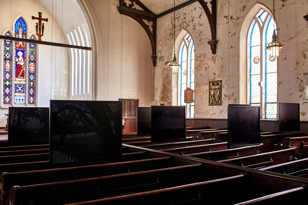 dawoud bey's large, nearly all black prints from his night coming tenderly, black, project, installed in 2018 just above the pews in the somewhat decaying st johns episcopal church in cleveland. the white walls are a bit flaky, but the stained glass behind the altar and the large arched windows bring in a lot of light. the black bey photos are almost illegible unless you get right up close. via front international