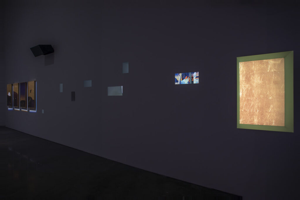 a wall in a dark gallery photographed at an angle with various rectangular forms along it, receding into the left distance. one group of four vertical rectangles are lit gold-ground icon style paintings; a series of apertures in the wall, most around painting height, fill the middle, and on the near right is either a monitor showing a painting or a spotlit gold ground painting. mark leckey installation view at gladstone gallery, nyc, nov 2024-feb 2025