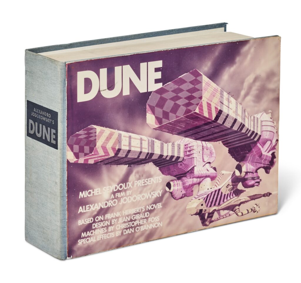 a copy of the jodorowsky dune bible photographed by christies at a slight angle to show the faded blue cloth spine, the 300 page thickness, and the pasted on color cover image with a hawk bodied spaceship with two star trek like nacelle things sticking out the top, except they're hollow like beveled paper towel tubes, and are maybe guns or something idk, the whole ship is painted with contrasting black and white strips and red and white checkerboards and chevrons, and the title dune is in fat non serif all caps, and a bunch of collaborators are listed below. this copy is number 4, and is for sale at christie's london in december 2024