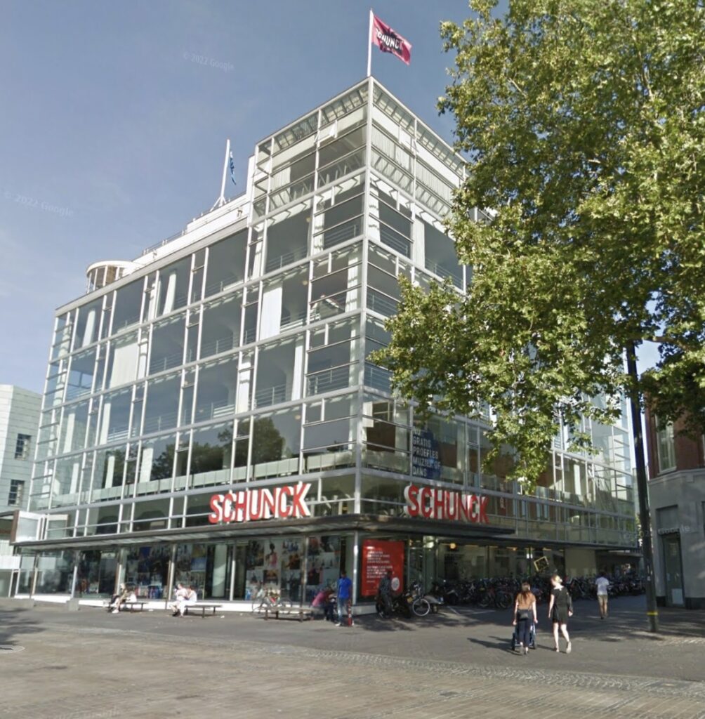 google streetview image from 2019 of schunk glass palace in heerlen, a color image of the all glass and steel skin of the eight story building with a red flag atop it, and a slightly larger tree in the right foreground. the building is now the town's cultural center and a music school.