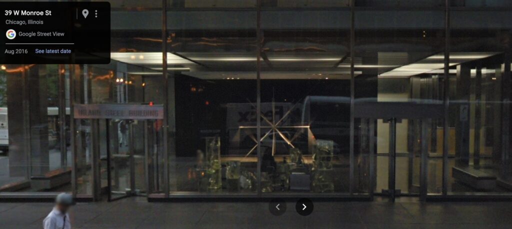 google streetview of chicago's inland steel building modernist steel and glass lobby, with richard lippold's eight pointed star sculpture dramatically lit from a suspended ceiling structure above it, and frank gehry's cast glass security desk sort of lost in the unlit middleground between the revolving doors. circa 2016