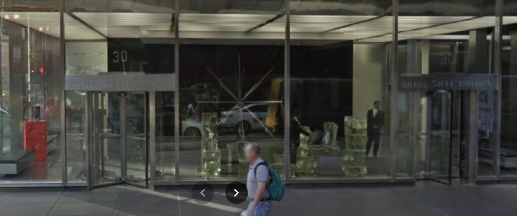 google streetview image of the lobby of the inland steel building in chicago with full unobstructed glass panels between steel mullions, richard lippold's eight pointed star shaped sculpture radiant 1 against the black background of the lobby wall, and frank gehry's cast glass blobs assembled into a security desk against the glass, between the two revolving doors. a bright red pedestal with perhaps a model of the building itself is against the left glass wall. circa 2017