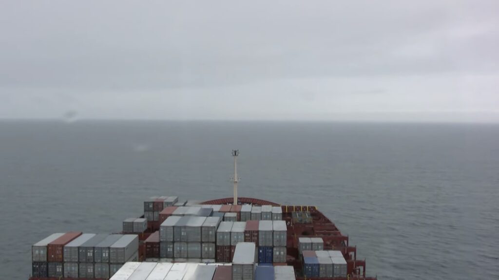 a screenshot of logistics, a 2012 film that lasts 37 days, shows the bow of a ship with containers stacked variously in five rows, on a grey sea and under a lighter grey cloudy sky. two rain drops on the camera housing distort the light slightly.