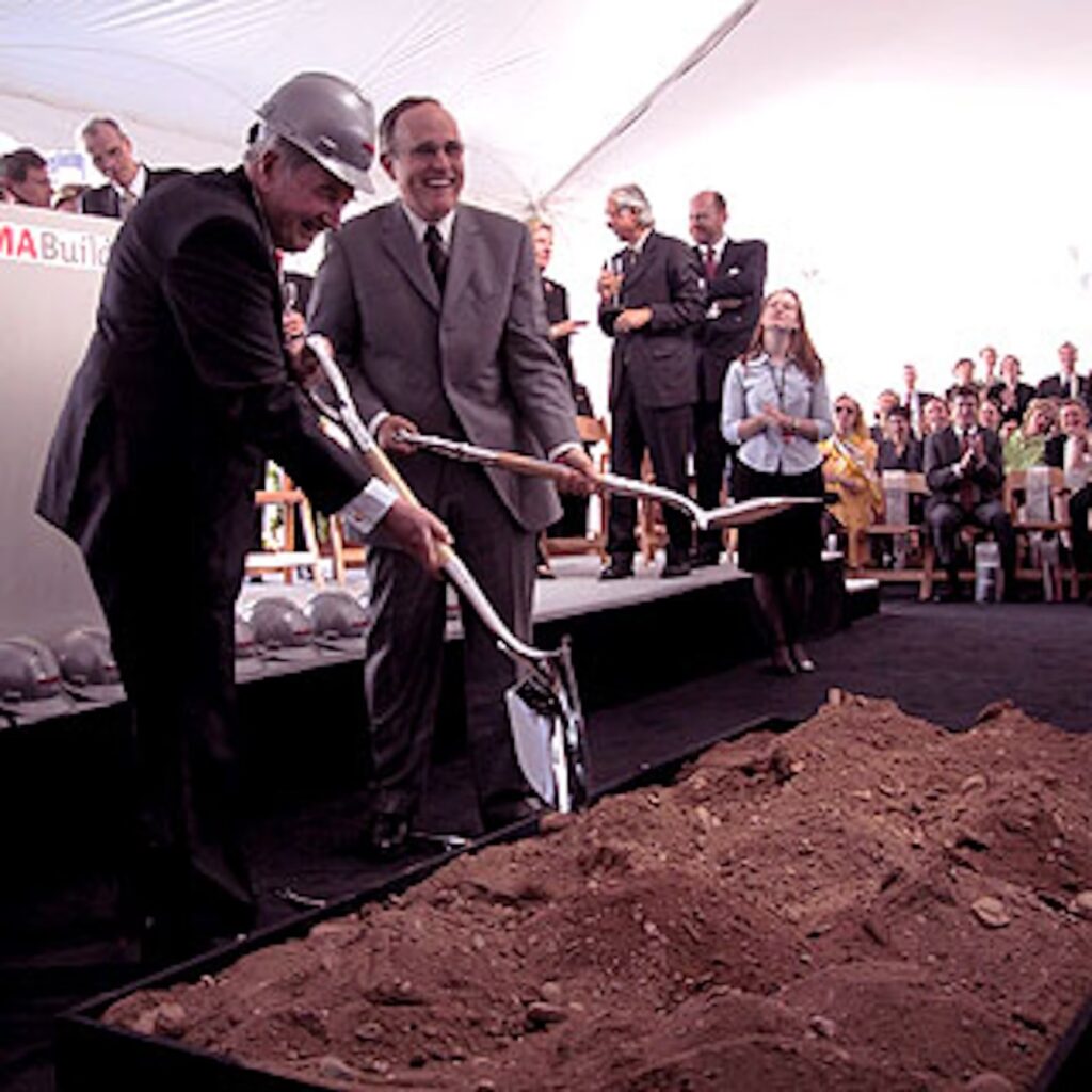 david rockefeller and rudy giuliani hold silver polished shovels and fake dig in some dirt for the groundbreaking of moma's yoshio taniguchi renovation. taniguchi and agnes gund and other folks stand on the dais behind, waiting their photo opp turn. the setting is a white tent, with some crowds visible in the background, applauding. image circa may 2001 via bizbash, a corporate party rental portal that is going strong in 2024, i guess