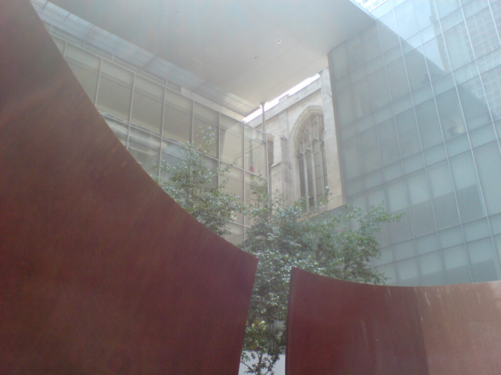 a gothic window of st thomas's church on fifth avenue is tightly framed by the glass walls and soaring white steel roof of the porch of yoshio taniguchi's moma, while a tree in the sculpture garden inhabits the middleground, and two curved plates of rusted corten steel of a richard serra torqued ellipse fill the left and lower section of the photo, taken from inside serra's sculpture in 2005