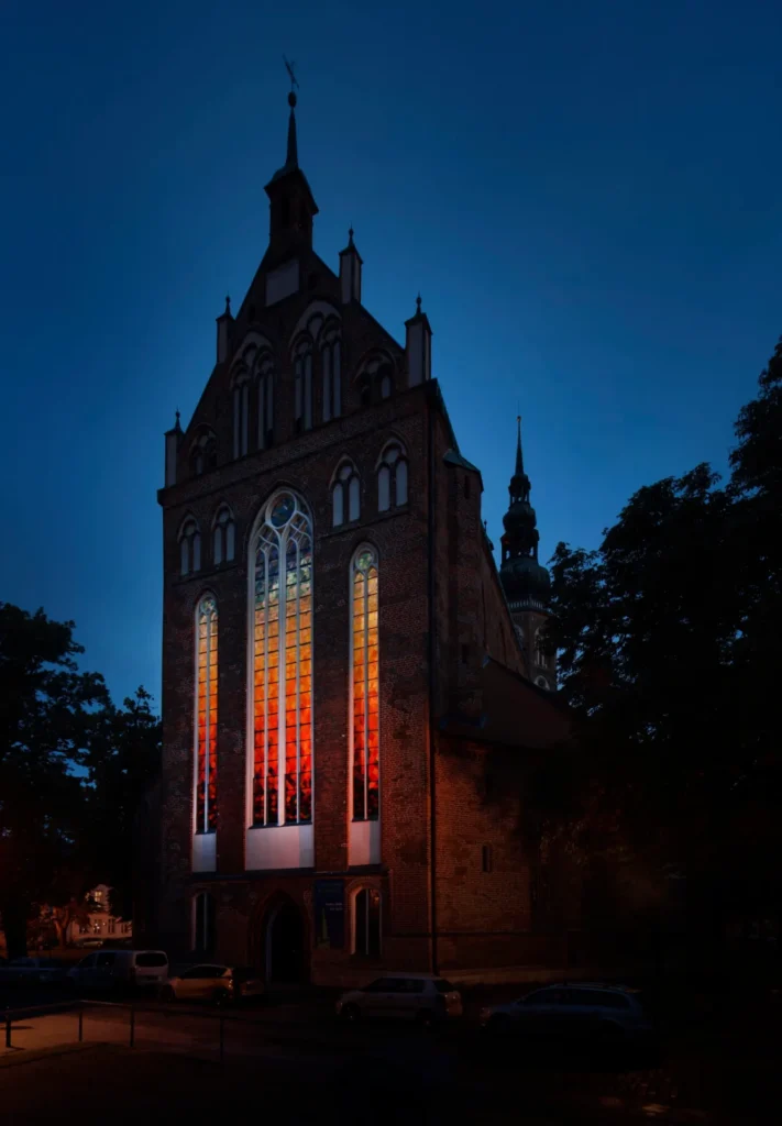 the east facade of a gothic brick cathedral in greifswald germany is dark against the deep blue sky of late afternoon or early evening, flanked by shadowy trees. the three tall, thin gothic windows with white tracery glow in a rainbow spectrum, dark red at the bottom to deep blue at the top, so mostly oranges and yellows in the middle, an installation by olafur eliasson