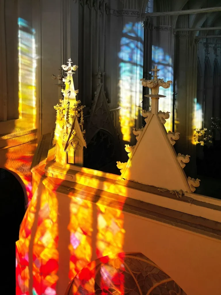 a closeup of ornately carved gothic finials on a wooden choir wall that are painted what seems to be white, with the larger space of the cathedral nave extending into the background, but the main feature of this photo is this architecture bathed, from bottom to top, in red, orange, yellow, and blue light, pouring in through the handblown glass window installation by olafur eliasson. at the st nicholas cathedral in greifswald germany.