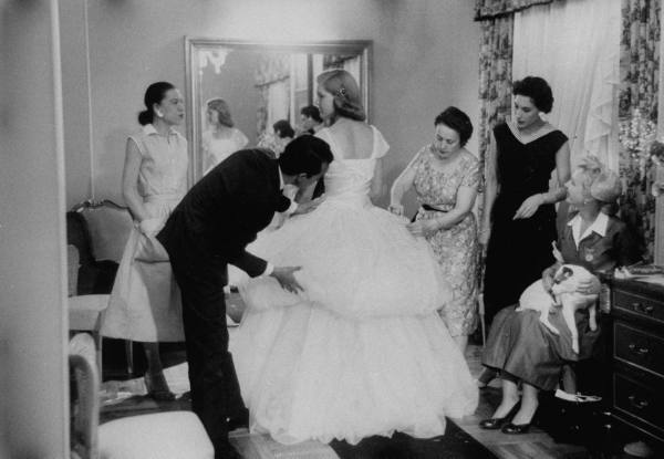 a black and white life magazine photo shows a dress fitting at the us embassy in madrid, where a young oscar de la renta in a black suit fluffs the tulle of a blonde 18 yr old white girl's white debutante gown, as her mother, two lady attendants, and the baroness de bodisco look on. photo credit; nina leen