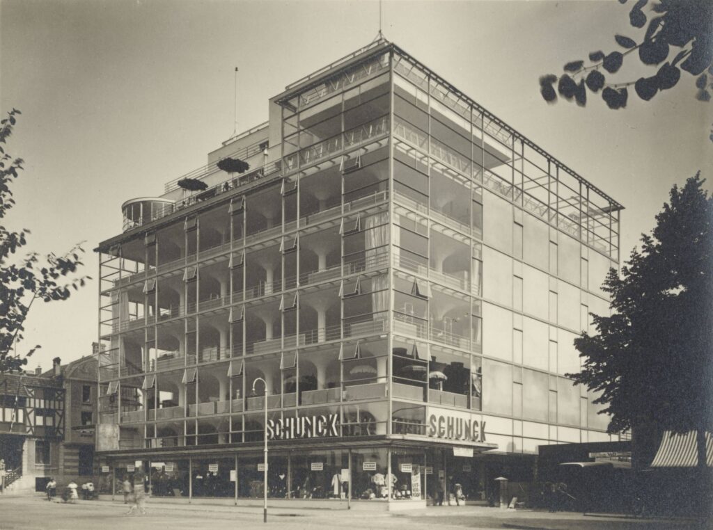 a black and white 1936 print of a photo by werner mantz of schunk department store in heerlen, the netherlands, shows an eight story square building encased in large sheets of glass curtain wall, which leaves the internal concrete slab and flared column structure completely visible and unobstructed. the plaza in front of the store is framed with trees along the image's edges. for sale at grisebach, a german auction house, in dec 2024