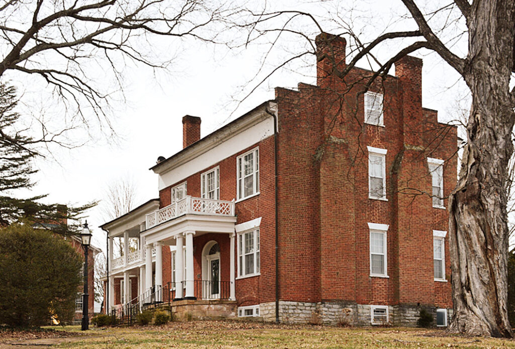 the reid white house, a 19th century red brick house in downtown lexington virgnia, is photographed from this oblique angle because just to the left, behind that hedge, is the post office, which is built on the house's front yard, and so there's no clear view of the house. it has two chimneys on each end, and a white columned porch. the double porch in the background was added later. via vlr