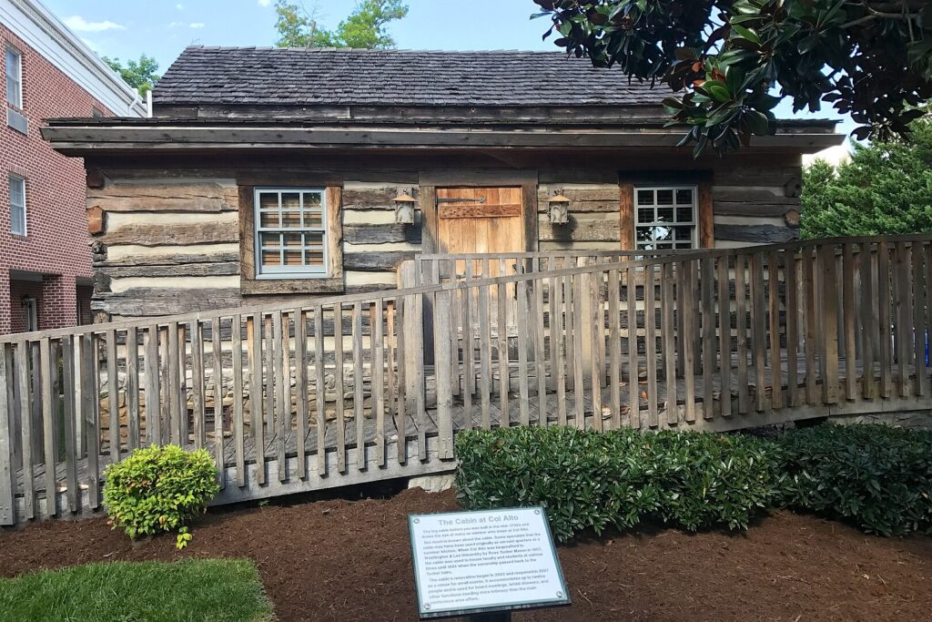 a circa 1700s log cabin with a deckwood wheelchair ramp in front of it, leading to a wood door flanked by lanterns and two small six-over-six windows, on the mulch and shrub landscaping in front of it is a plaque explaining how no one knows what this cabin was for, maybe it was a summer kitchen or quote servant quarters, but it sits on the grounds of a virginia estate that is now a hampton inn. a modern annex of the hotel, in brick, pokes out of the left side of the photo, while the parking lot right up against it has been cropped out.