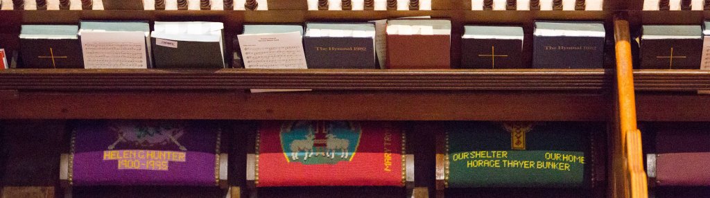 a tight photo of the back of a pew at the trinity church boston has a row of prayerbooks and hymnals along the top, and three kneelers—movable cushion-topped stools for worshipers to kneel on during the service—covered in needlepoint of various designs. each is dedicated to someone, perhaps someone who has died; the one on the left is purple and gold and has the name of a lady who died at 95. the center one is red with some sheep in a shield-shaped field, and the one on the right is dark green with a cross and lettering in gold that reads our shelter our home. image via trinity church boston dot org