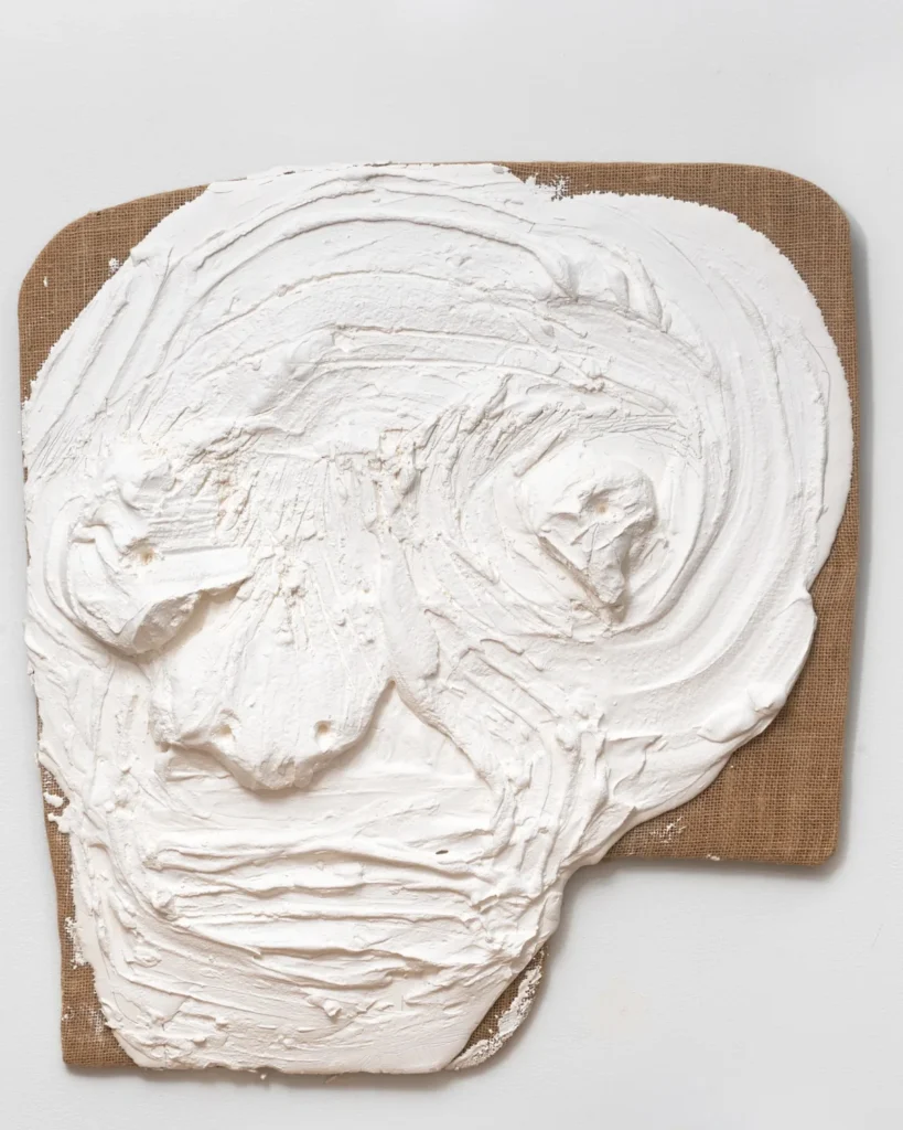 a swirly frosting-like surface of white plaster on a burlap wrapped panel shows the finger marks of nicole eisenman, the artist who sculpted a rough, slightly abstracted face out of it. from a group titled understudies, on view at anton kern gallery in early 2025