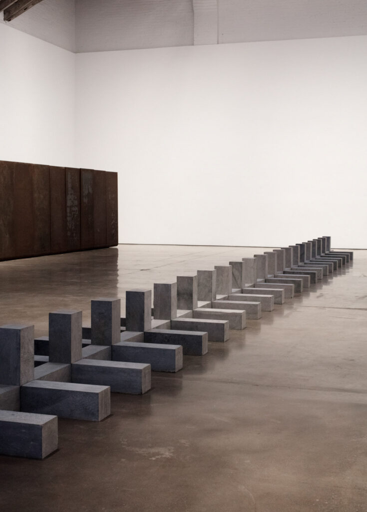 two sculpures by carl andre, who got off for killing his wife, installed in the paula cooper gallery in january 2025. there's a bent metal thing on the back wall that's not important, and 101 belgian block stones arranged like half a zipper running frrom the lower left corner toward the back right wall. this is one of the works andre made in and showed only in europe in the time between the murder and his indictment in 1985, and his trial in 1988. photo: camille drury