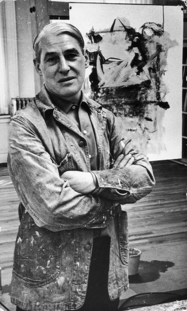 a waist-up portrait of willem de kooning standing confidently with his arms folded, wearing a paint splattered denim work coat and a smirk, in a photograph taken in his soho loft studio in march 1962 by fred mcdarrah, which, getty images forces a caption for, but which came from village preservation dot org, where fred's estate has donated some fundraising prints