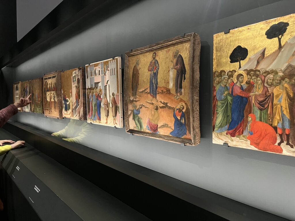 an oblique image of all eight back predella panels of duccio's maesta altarpiece, painted in 1308-1311. closest on the right are christ raising lazarus, surrounded by a crowd, with a red robed figure at his feet; then the transfiguration, christ at the center of five disciples at a distance on a mountaintop; and christ healing the blind man, set in a perspectival city plaza, with christ in profile and a crowd behind him on the left. these eight panels are installed in a row in a dark vitrine at the metropolitan museum in winter 2024-25, the first time they've been reunited since being chopped up and sold for parts