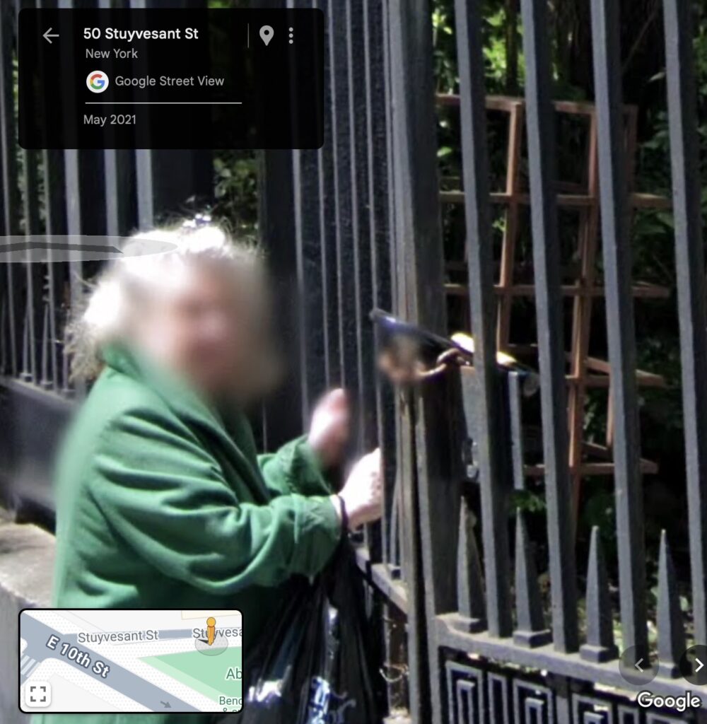 a detail from a google streetview screencap from may 2021, where an elderly white woman in green coat has her hands on the chained wrought iron gate of st marks church in the bowery. gordon matta clark's rosebush sculpture is behind the gate, an unpainted rust color