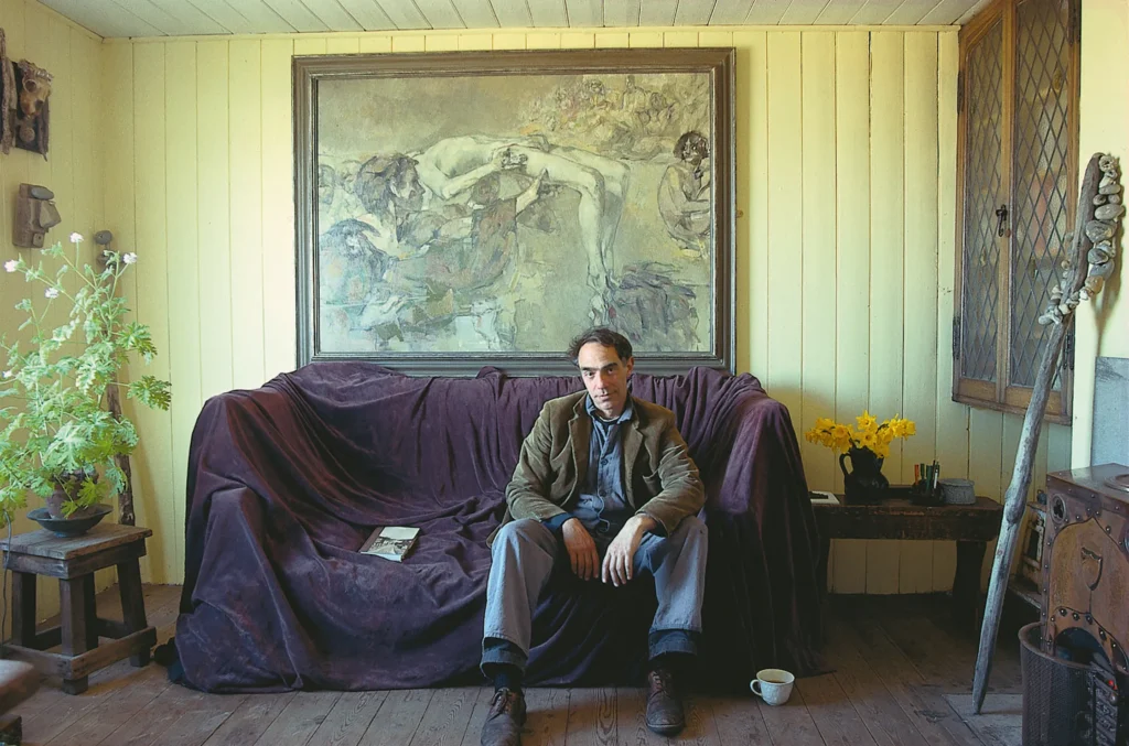 filmmaker derek jarman sits on a sofa covered with purple velvet in the yellow painted living room of his cottage in 1989. he is white with black hair, wearing a denim shirt under a mushroom colored jacket. a book sits next to him on the sofa, and a large pale toned painting of a reclined nude white male figure with another male figure in a tunic in front of it, and sketched out crowds in the periphery. in the room, a plant sits on a low stool at the left, diamond pane cabinet windows inset into the wall on the right have a piece of driftwood studded with pebbles or something. the photo was taken for world of interiors magazine by john vere brown, and they have run it three times so far.