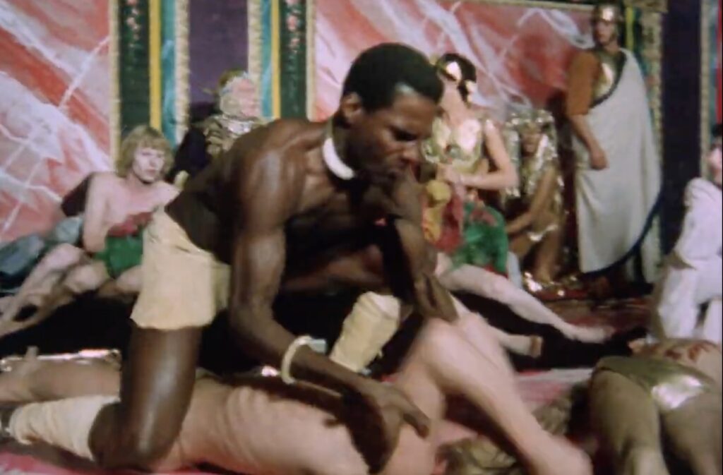 a screenshot of the opening scene of derek jarman's sebastiane in which a black slave in a loincloth has killed one pink twink in a lamé bikini on the edge of a colorfully painted dais, and is now manhandling another one, just before he kills him too. figures in approximated roman dress are visible in the background, as is a handpainted pink and green marbled backdrop