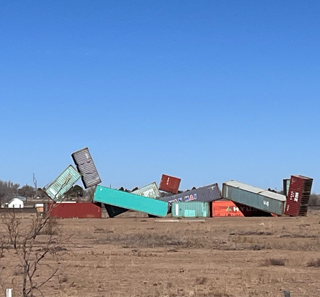 under a cloudless blue sky, twelve shipping containers of various lengths are arranged in precarious-seeming angles to resemble a human figure with its legs crossed and its head propped on one hand in a monumental sculpture by matt johnson titled, sleeping figure. just in case you didn't know it was sleeping, or a figure, though the artist apparently painted sleepy eyes and a closed mouth on what's supposed to be the head. hopefully someone will buff those out soon. this photo of the sculpture installed in marfa texas was posted on bluesky on jan 1, 2025, a good beginning to a cursed year, by stacey burns, wentrogue.bsky.social.