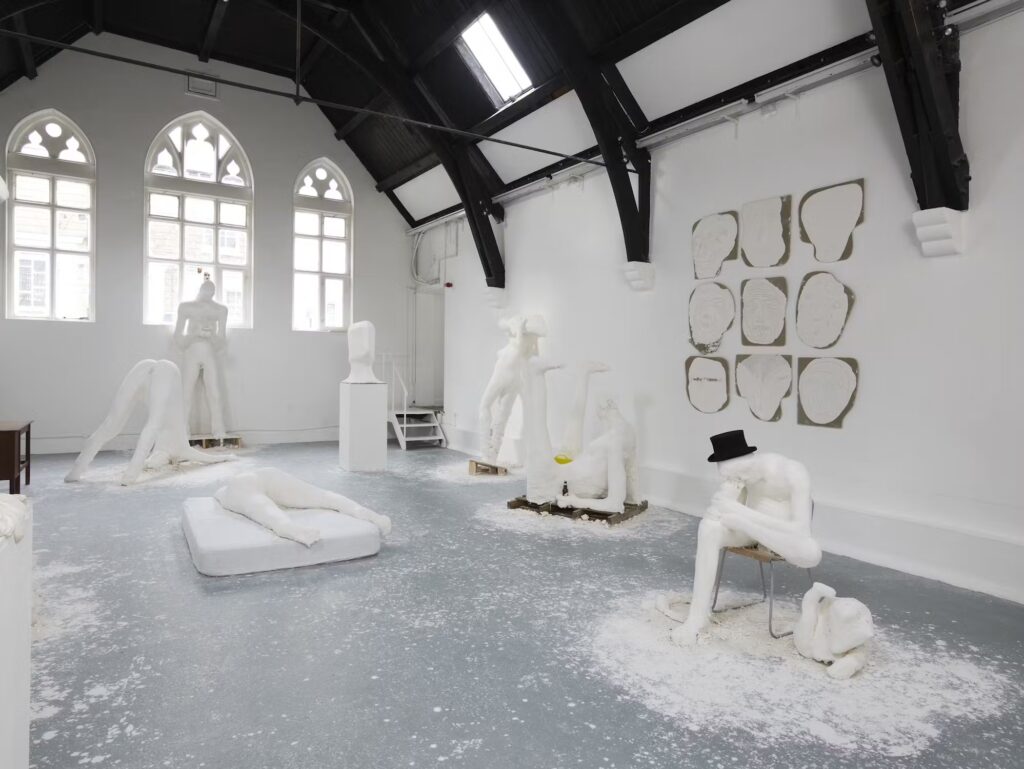 a gallery space in a repurposed church-like hall in london with a high beamed ceiling and three gothic pointed windows on the end wall has white walls and a light grey floor, which is splattered with plaster, particularly around the roughly made, awkardly shaped and posed plaster figures in the space, a clear sign the artworks were made there. on the wall is a 3x3 grid of plaster headlike shapes, flat on burlap wrapped board, some more obviously faces than others. a 2012 show by nicole eisenman at studio voltaire.