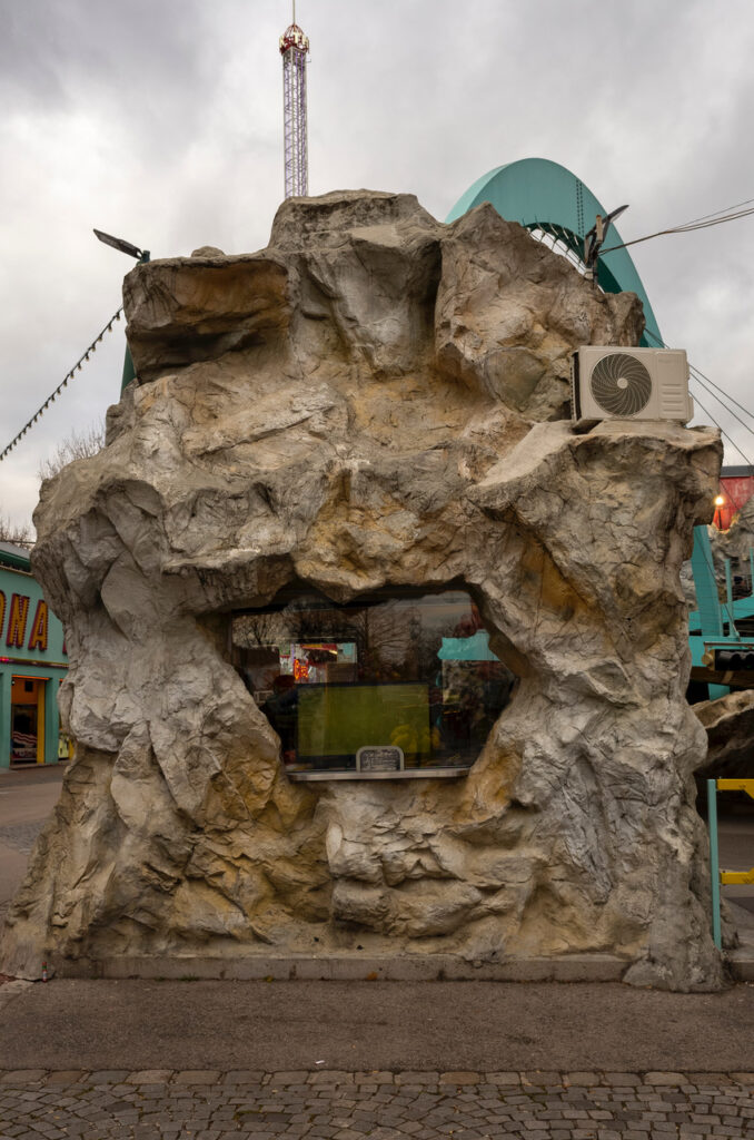 a freestanding ticket booth at wurstelprater, an amusement park in vienna, is built to resemble a graggy brownish grey cliff two stories tall, except it rests on a plinth of stone or concrete, which the concrete fake rock appears to overlap, like an overgrown tree root. and there is a wide plate glass window with an irregular edge and a level shelf, which contrasts with the seeming natural wildness of the rock. and on the upper right is an air conditioning compressor on its own ledge cut into the fake rock, an even starker contrast of nature and machine than the window. power lines, the tops of various rides behind the booth, etc. poke up from the top. the bottom edge foreground is a neat cobblestone 