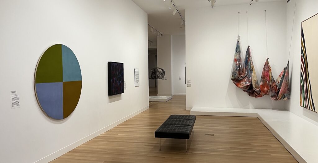 a narrow white cube gallery at the smithsonian american art museum with wood floors and a black mies bench at the center, features all dc artists: from left, a round painting divided into muted 60s color quadrants, the only public painting by mary pinchot meyer; and a dark atmospheric nebula-filled painting by kenneth young are on one wall. on the right, with a plinth to keep people back, a paint soaked canvas by sam gilliam hangs in swags from four chains in the ceiling, and next to it, the poured rainbow corner of a wider morris louis painting. 10/10 no notes