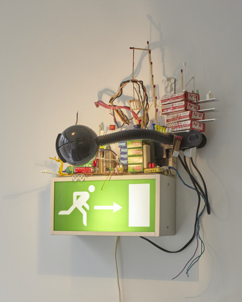 sarah sze's sculpture from 1997 is an accumulation of batteries, wires, tic tacs, aspirin, packs of gum, little boxes of raisins, and a clip on reading lap, all hot glued atop a green, backlit emergency exit sign of the type seen in france. now in the moma collection.