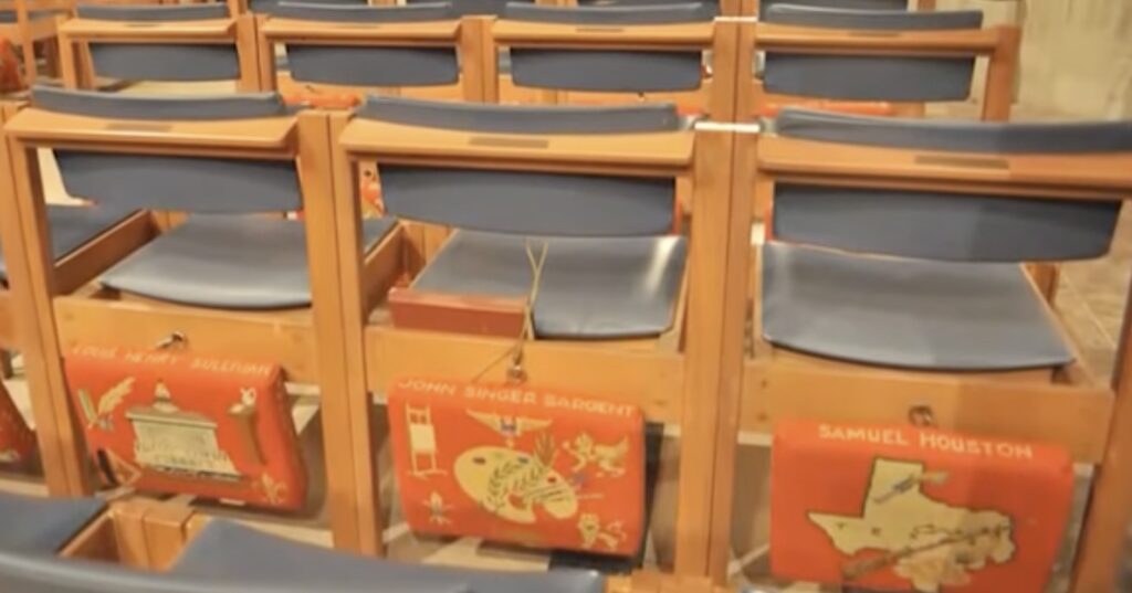 a sreenshot depicting the backs of three wooden pew seats at the national cathedral, with blue seat cushions, and hanging behind each chair, a red needlepoint upholstered kneeling cushion for the use of the worshipers on the row behind. the edge of the kneelers are decorated with the name of some historically significant figure, and the face of the kneeler depicts several corny symbols to reference that person's life or accomplishments. so on the right is sam houston, for example, and the state of texas. in the middle is john singer sargent and a palette, an easel, and an american eagle crest for some reason? on the right, who cares you get the idea. it goes on and on