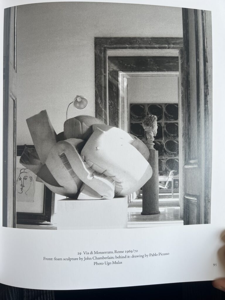 photo of a page in homes & studios of a 1969 or 1970 photo by ugo mulas of cy twombly's rome palazzo. through the darkly veined marble doorway is a roman bust on a too thin pedestal and behind it, the repetitive grid of silkscreened cans of andy warhol's tuna disaster painting. in the foreground, a massive foam sculpture by john chamberlain on a pedestal partially blocks a framed line drawing by picasso, according to the caption. it looks like a 1943 drawing, but could be a copy made by twombly, which is a thing he did sometimes.