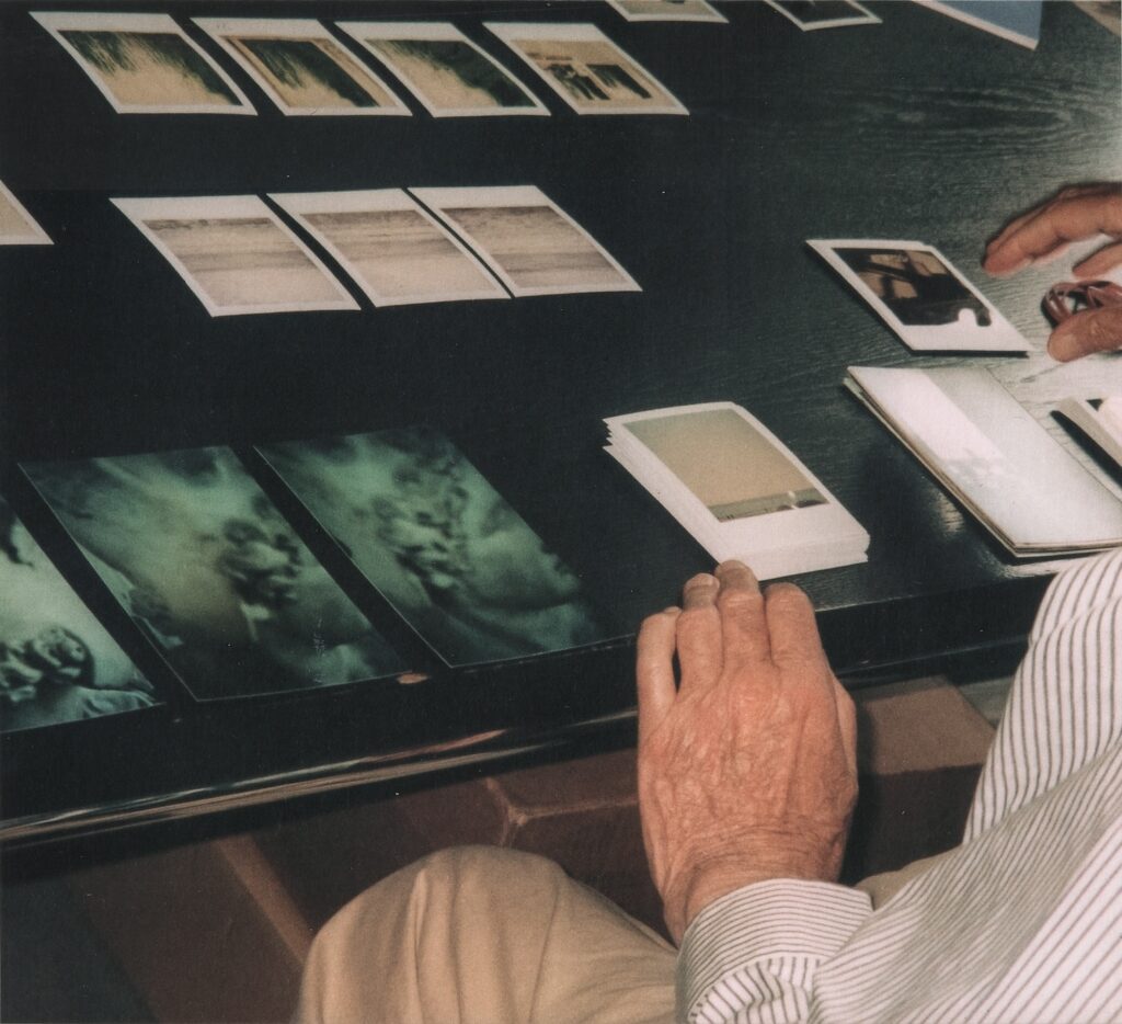 a 2010 photo by nicola del roscio over cy twombly's left shoulder as the artist sits in khakis and a red and white pinstripe shirt at a black tabletop. his aged hands move toward stacks and rows of polaroid photos arranged on the table. closest to the edge are three closeup details of a classical sculpture of a male head. two rows of polaroids depict variations of light and dark shadows and lines, perhaps details from some painting or other. via cy twombly dot org