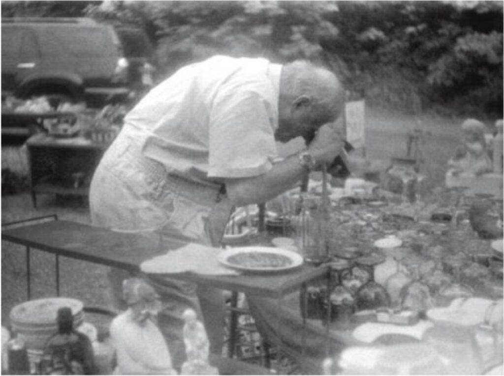 cy twombly at an outdoor rummage sale, bent over, a table full of glassware, possibly looking in a microscope, possibly photographing something with a polaroid camera, in a photo taken by sally mann, and published in her memoir hold still